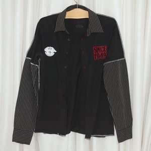 May include: A black and white striped long-sleeved shirt with a black button-up front. The shirt has a patch on the left side that says "Detroit" and a red patch on the right side that says "Since 1949".