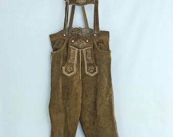 May include: Brown leather lederhosen with suspenders, hanging on a wooden hanger. The traditional Bavarian pants feature decorative embroidery on the front and suspenders. The pants have pockets and tie closures at the ankles.