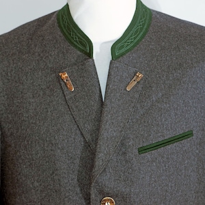 May include: A grey wool jacket with a green collar and pocket trim. The jacket features two decorative metal clasps and a gold button. The collar has an embroidered design. This traditional jacket is suitable for formal occasions.