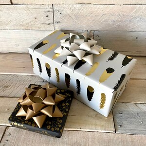 May include: Two wrapped gifts with decorative bows. One gift is wrapped in white paper with gold and black feather patterns. The other gift is wrapped in black paper with gold floral patterns. Both gifts have large bows.