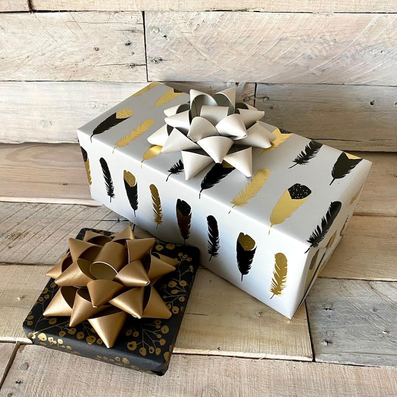May include: Two wrapped gifts with decorative bows. One gift is wrapped in white paper with gold and black feather patterns. The other gift is wrapped in black paper with gold floral patterns. Both gifts have large bows.
