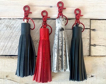 May include: Four leather tassel keychains with red metal clasps. The tassels are black, red, white and gray, and are hanging from a wooden surface.