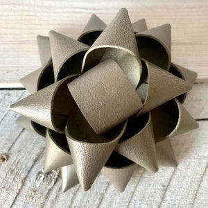May include: A decorative, star-shaped bow in a metallic gray color. The bow is made of overlapping, cone-shaped pieces, creating a layered effect. The bow is likely made of a faux leather material, and is suitable for gift wrapping or craft projects.