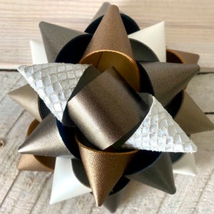 May include: A multi-layered, star-shaped hair bow. The bow features various textures and colors, including white, silver, gold, and dark gray. The bow is made of leather-like material and is suitable for various occasions.