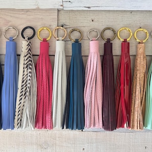 Op de afbeelding: Een collectie van elf decoratieve kwastjes in diverse kleuren en texturen, waaronder zwart, blauw, dierenprint, roze, wit, donkerblauw, lichtroze, donkerrood, felrood, beige kurk en mintgroen, elk bevestigd aan een ronde metalen ring.