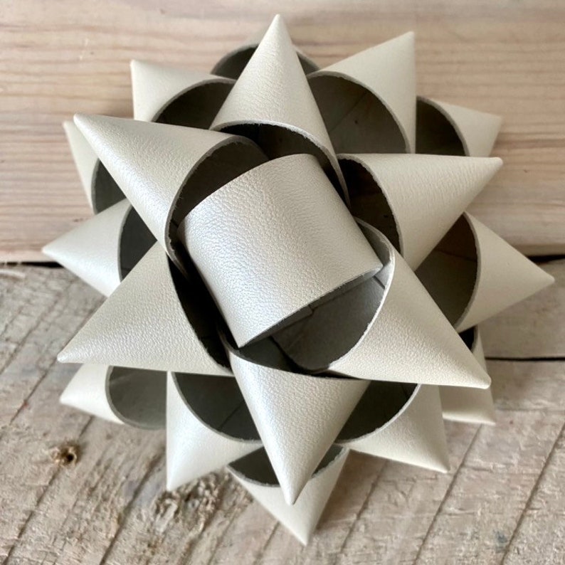 May include: A decorative, off-white bow made of textured material. The bow is shaped like a starburst, with multiple cone-shaped elements arranged in a circular pattern. The bow is placed on a light-colored wooden surface.