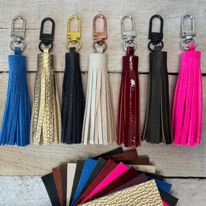 May include: A collection of keychains with colorful leather tassels. The keychains have silver, black, and gold-toned hardware. Tassels are blue, gold, black, cream, burgundy, dark brown, and pink. A fan of leather swatches in various colors is below.