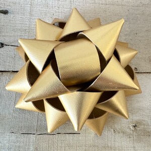 May include: A gold-colored decorative bow with a starburst design. The bow is made of a metallic material and has a textured surface. The bow is sitting on a light-colored wooden surface.