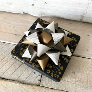 May include: A square gift box wrapped in black paper with a gold floral pattern. A large bow made of white, silver, gold, and brown faux leather is on top of the box. The bow has a textured, layered design.