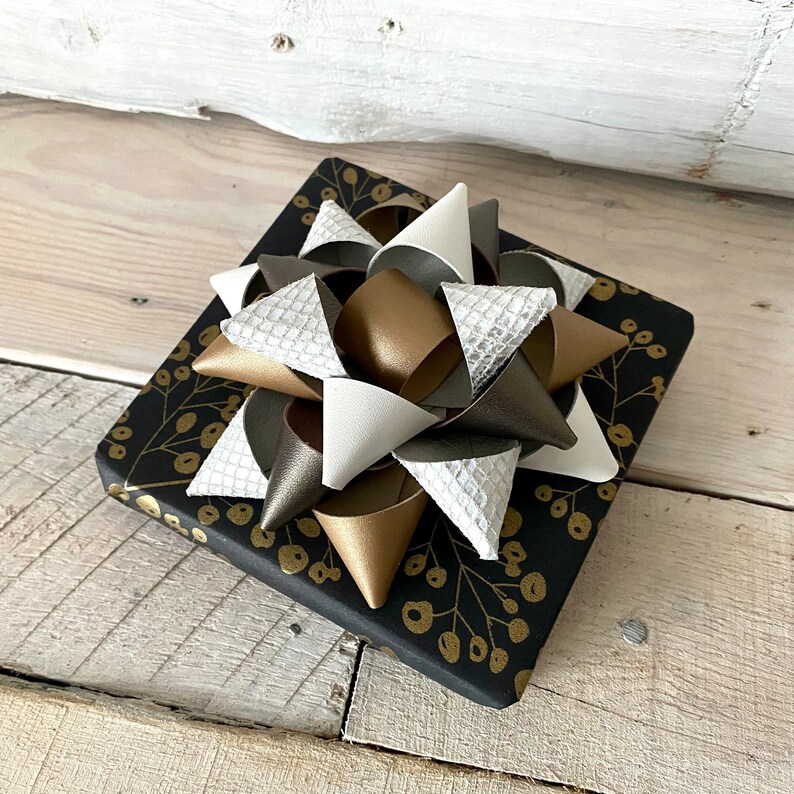 May include: A square gift box wrapped in black paper with a gold floral pattern. A large bow made of white, silver, gold, and brown faux leather is on top of the box. The bow has a textured, layered design.