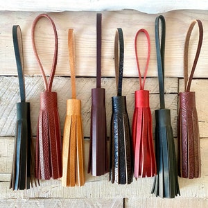 May include: Assortment of eight leather tassels in various colours, including black, brown, tan, and red. The tassels are all approximately the same size and have a loop at the top for hanging.