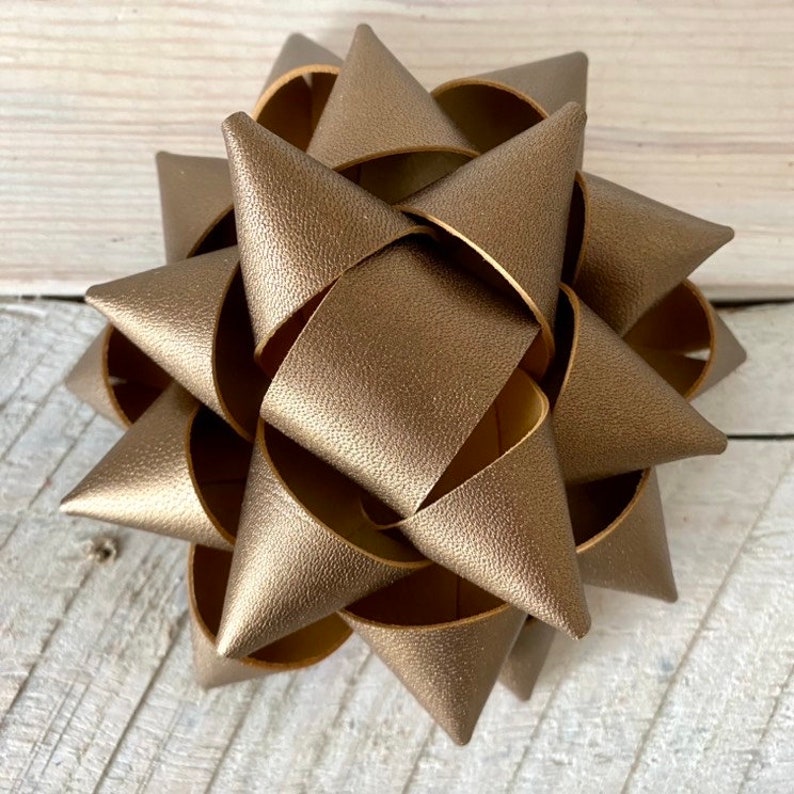 May include: A metallic gold-colored decorative bow with a starburst design. The bow is made of overlapping, textured material, creating a three-dimensional effect. The bow is likely used for gift wrapping or other decorative purposes.