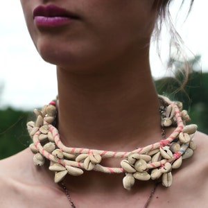 Op de afbeelding: Een beige schelpenketting met roze en witte touwaccenten. De ketting heeft meerdere touwstrengen versierd met talrijke schelpen. De sieraden worden om de nek gedragen.
