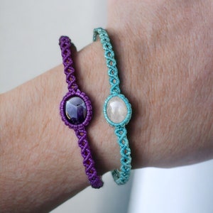Puede incluir: Dos pulseras con cordones anudados y detalles de piedras preciosas. La pulsera de la izquierda tiene un cordón morado y una piedra preciosa morado oscuro. La pulsera de la derecha tiene un cordón turquesa y una piedra preciosa blanca.