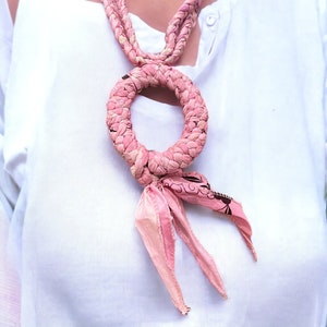 Op de afbeelding: Een roze stoffen ketting met een gevlochten touwontwerp en een ronde hanger. De hanger is vastgebonden met een roze lint met een bloemenpatroon.