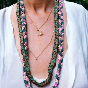 Op de afbeelding: Een lange, gevlochten ketting gemaakt van stof in tinten groen, bruin, roze en wit. De ketting wordt gedragen over een gouden ketting met een klein hangertje.