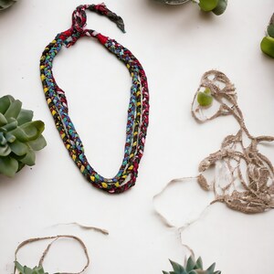 Op de afbeelding: Een veelkleurige gevlochten ketting gemaakt van stof met een rood, blauw en geel kleurenschema. De ketting ligt op een wit oppervlak met verschillende kleine groene vetplanten in potten.