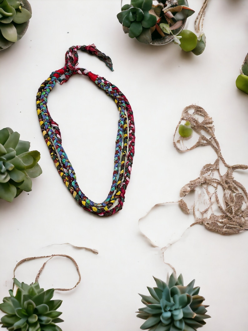 Op de afbeelding: Een veelkleurige gevlochten ketting gemaakt van stof met een rood, blauw en geel kleurenschema. De ketting ligt op een wit oppervlak met verschillende kleine groene vetplanten in potten.