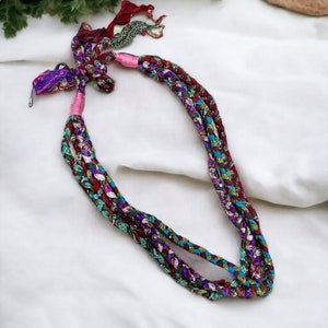 Op de afbeelding: Een veelkleurige gevlochten ketting gemaakt van stoffen restjes. De ketting is vastgebonden met een roze lint en heeft een metalen sluiting.
