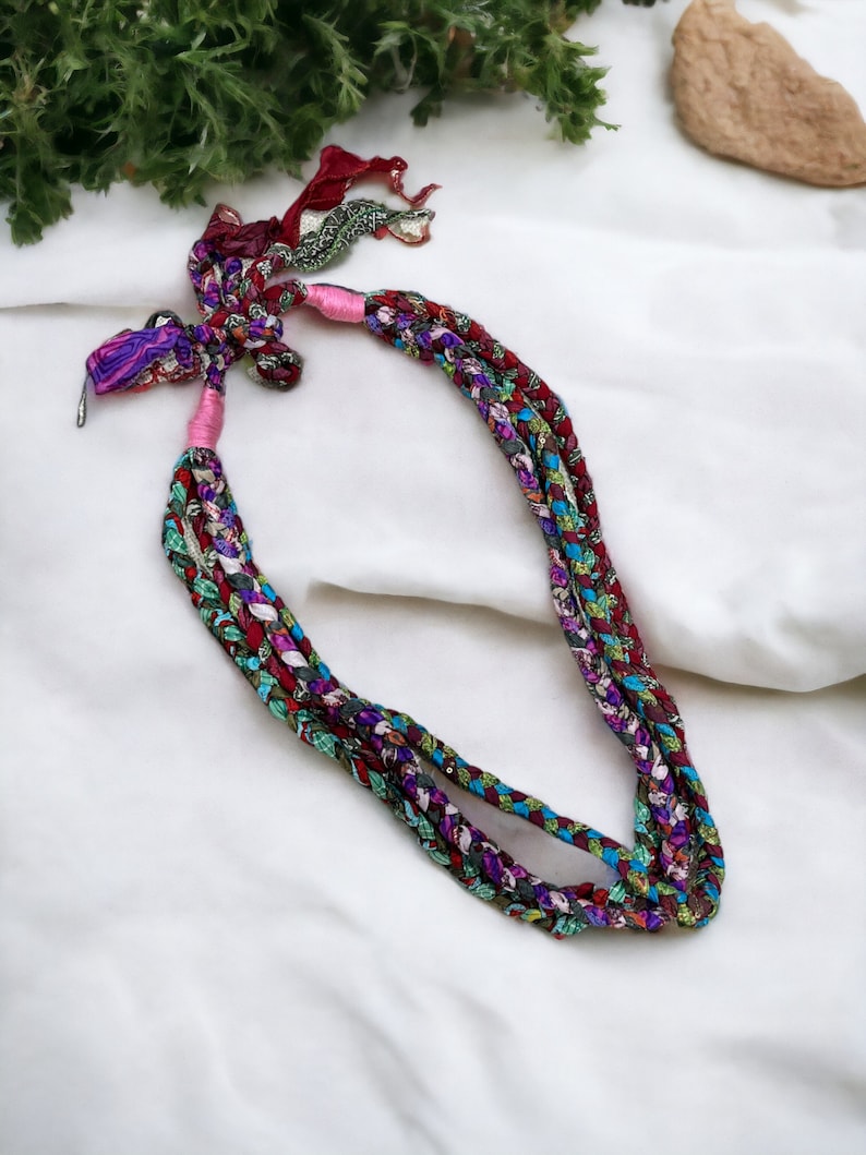 Op de afbeelding: Een veelkleurige gevlochten ketting gemaakt van stoffen restjes. De ketting is vastgebonden met een roze lint en heeft een metalen sluiting.