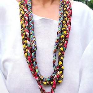 Op de afbeelding: Een veelkleurige gevlochten ketting gemaakt van stofresten. De ketting is gelaagd en heeft tinten rood, groen, geel, blauw en bruin.