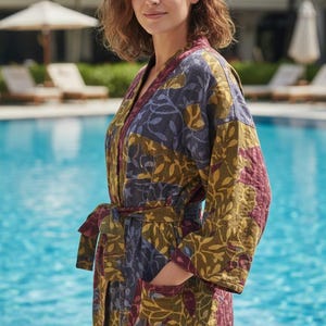 Pode incluir: Robe estampada com cinto, em verde oliva, bordô e azul. O robe tem um desenho de folhas e é usado por uma pessoa perto de uma piscina. O robe tem bolsos e mangas compridas.