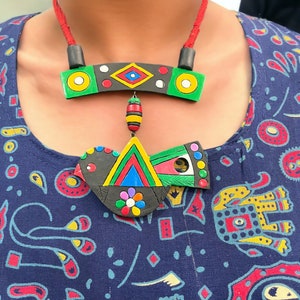 Op de afbeelding: Een kleurrijke, handgemaakte ketting met een geometrisch ontwerp. De ketting heeft een zwarte basis met groene, gele, rode en blauwe accenten. De hanger hangt aan een rood koord. De ketting wordt gedragen over een blauwe top met patroon.