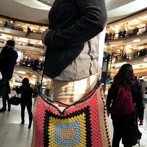 May include: A large, colorful crocheted tote bag with a square pattern in pink, yellow, blue, and black. The bag is worn over the shoulder of a person in a shopping mall.
