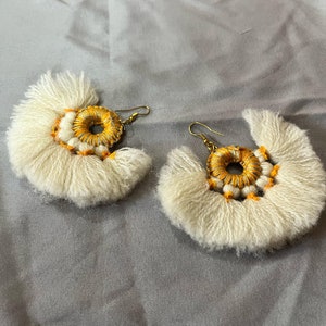 May include: A pair of white tassel earrings with gold and orange accents. The earrings are in the shape of a crescent moon and have a circular design in the center.