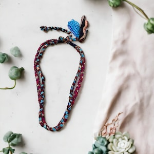 Op de afbeelding: Een gevlochten ketting met een blauw en rood patroon en een kleine blauwe en witte hanger. De ketting ligt op een wit oppervlak met groene planten op de achtergrond.