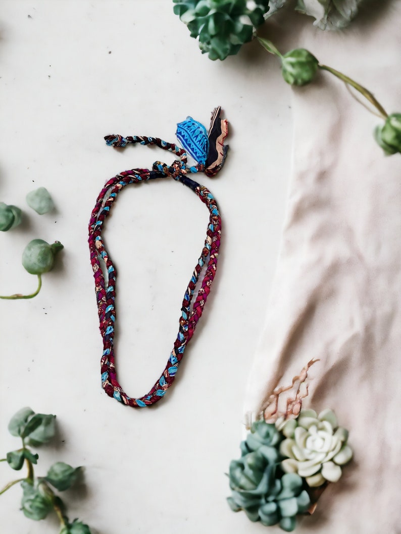 Op de afbeelding: Een gevlochten ketting met een blauw en rood patroon en een kleine blauwe en witte hanger. De ketting ligt op een wit oppervlak met groene planten op de achtergrond.