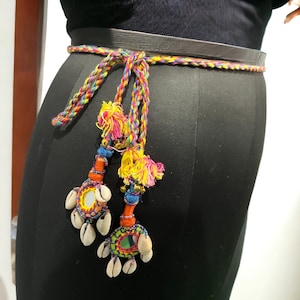 Op de afbeelding: Een kleurrijke gevlochten riem met kwastjes met kleine spiegels en schelpen. De kwastjes zijn gemaakt van kleurrijk garen en kralen.