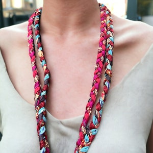 Op de afbeelding: Een lange, veelkleurige gevlochten ketting gemaakt van stof. De ketting is gemaakt van rode, blauwe, bruine en witte stof.