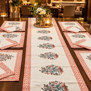 May include: A white table runner and matching placemats with napkins, all featuring a repeating floral motif in teal and rust orange. The fabric pieces have a decorative border in a geometric pattern of the same rust orange color, arranged on a table.