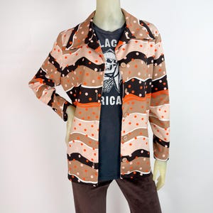 Può includere: Giacca vintage con bottoni e motivo retrò. La giacca presenta strisce orizzontali nei toni del marrone, nero e beige, accentate da pois arancioni e bianchi. È indossata sopra una maglietta grigio scuro con una grafica a teschio e la scritta "LAG RICA".