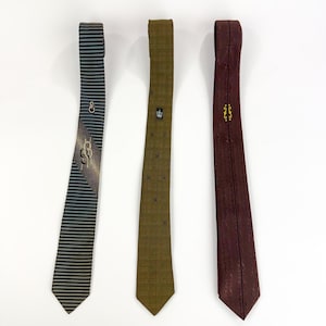 Op de afbeelding: Drie vintage stropdassen worden getoond tegen een witte achtergrond. De stropdassen zijn in tinten groen, bordeaux en zwart. Eén stropdas heeft een streepjespatroon met een gouden design, een andere heeft een kroonembleem en de derde heeft een textuurpatroon.