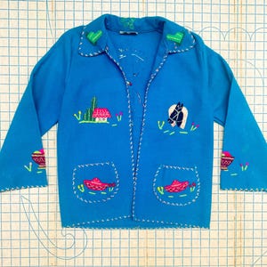Puede incluir: Cárdigan bordado azul turquesa con temática del suroeste. La chaqueta presenta bordados de cactus, un caballo y un sombrero. Los puños y el cuello tienen costuras decorativas. Los bolsillos están delineados con costuras blancas.
