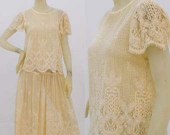 May include: A two-piece outfit made of delicate cream-coloured lace. The top has short sleeves and a round neckline. The skirt is knee-length and has a gathered waist.