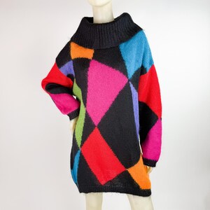 Op de afbeelding: Een jurk met lange mouwen en een zwarte coltrui en een geometrisch patroon. Het patroon heeft grote, kleurrijke ruitvormen in rood, roze, blauw, oranje, groen en paars. De jurk wordt op een mannequin getoond.