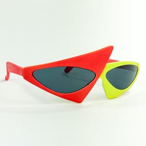 May include: Asymmetrical sunglasses featuring a large, triangular red frame on the left side and a smaller, rounded lime green frame on the right side. The lenses are dark grey and tinted, set within a bold, multi-coloured plastic frame structure.