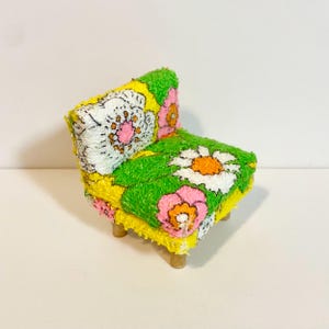 May include: A miniature armchair featuring a vibrant yellow, green, and white floral pattern with pink and orange accents. The chair has a textured, fuzzy surface and is supported by four small, light-colored wooden legs, creating a compact seat.