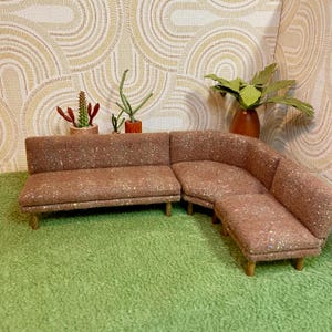 May include: A miniature mid-century modern sectional sofa in a speckled brown fabric. The sofa has a two-seat section, a corner piece, and an ottoman. The sofa sits on short wooden legs. Decorative plants and a patterned wallpaper are in the background.