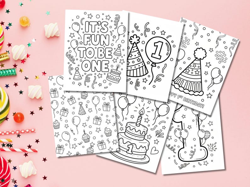 May include: A set of black and white birthday-themed colouring pages. The designs include birthday cakes, balloons, party hats, and the phrases "IT'S FUN TO BE ONE" and "HAPPY 1ST BIRTHDAY!". The pages are arranged on a pink surface.