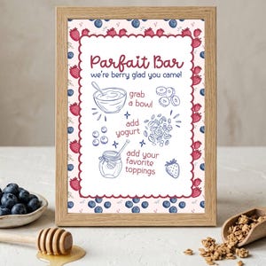 May include: A framed sign with the text "Parfait Bar" and instructions. The sign is decorated with strawberries and blueberries. A bowl of blueberries, a honey dipper, and granola are in the foreground.