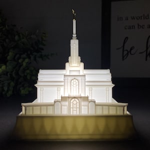 May include: A white 3D printed model of a temple with a gold spire. The model is lit from within, creating a warm glow. The model is sitting on a white base.