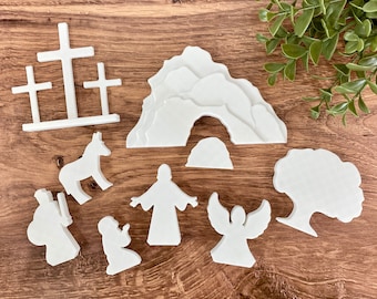 May include: White wooden cutout figures depicting the Easter story. The figures include a cross, a tomb, an angel, a donkey, Jesus, and two people.
