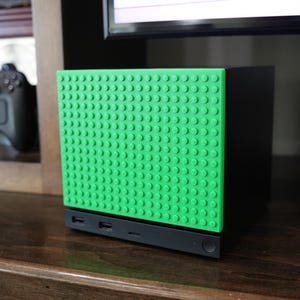 May include: A black gaming console featuring a bright green, textured front panel with a grid of raised circular studs. The base of the console is black and includes two USB ports, a small horizontal slot, and a circular power button.