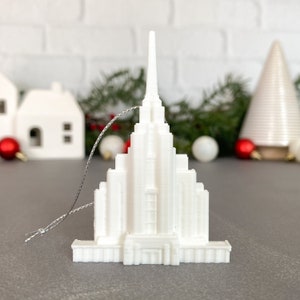 May include: White 3D printed ornament shaped like a church building with a pointed spire. The ornament has a silver string attached for hanging.