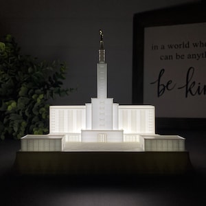 Può includere: Un modello bianco di un tempio stampato in 3D con una guglia e una piccola figura in cima. Il modello è illuminato dal basso, proiettando un bagliore caldo. Il modello è posto su una superficie scura.