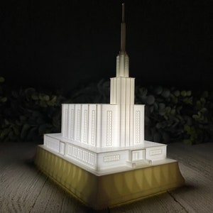 May include: A white architectural model of a temple with a tall spire, illuminated from within. The model sits on a gold-colored base, showcasing intricate details of the building's design. The background is dark, with hints of greenery.